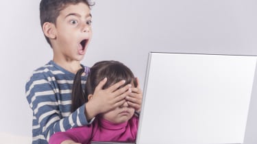 Two children using a computer with the older one cover the eyes of the younger one with an expression of shock