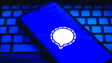 A smartphone sat on top of a white keyboard with the Signal app logo superimposed onto the phone&#039;s display - an image all set in dark blue and black lighting